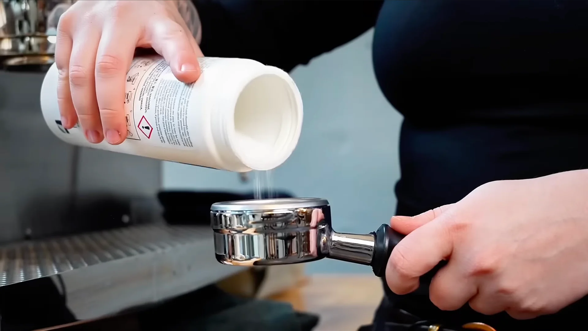 Staff member holding a portafilter filled with cleaning powder in front of a commercial espresso machine during routine maintenance