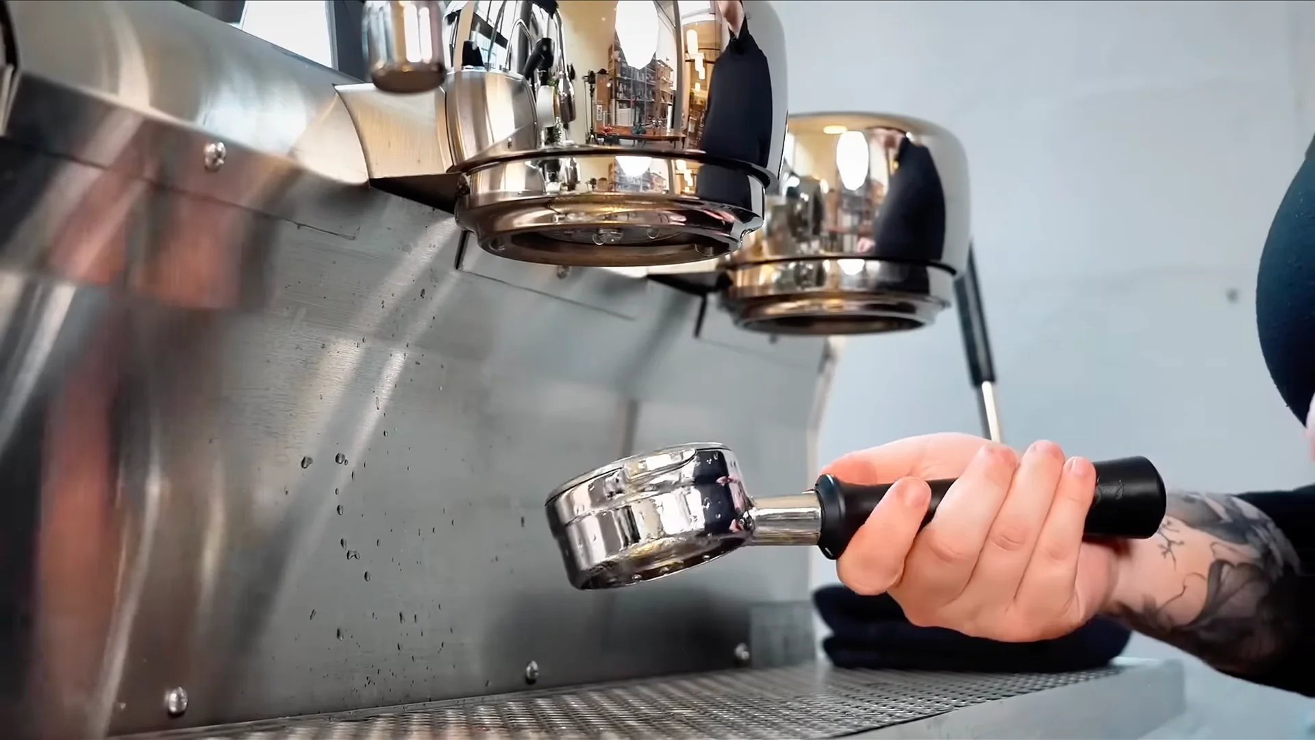 Barista wearing gloves cleaning an espresso machine steam wand in a measuring jug, with cleaning solution, cloth, and brush on a café counter